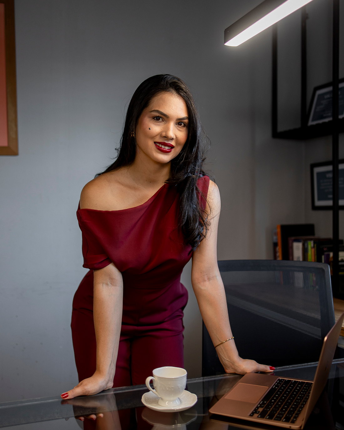 Dra. Elaine Pereira sorrindo no escritório, de vestido vinho, apoiada na mesa